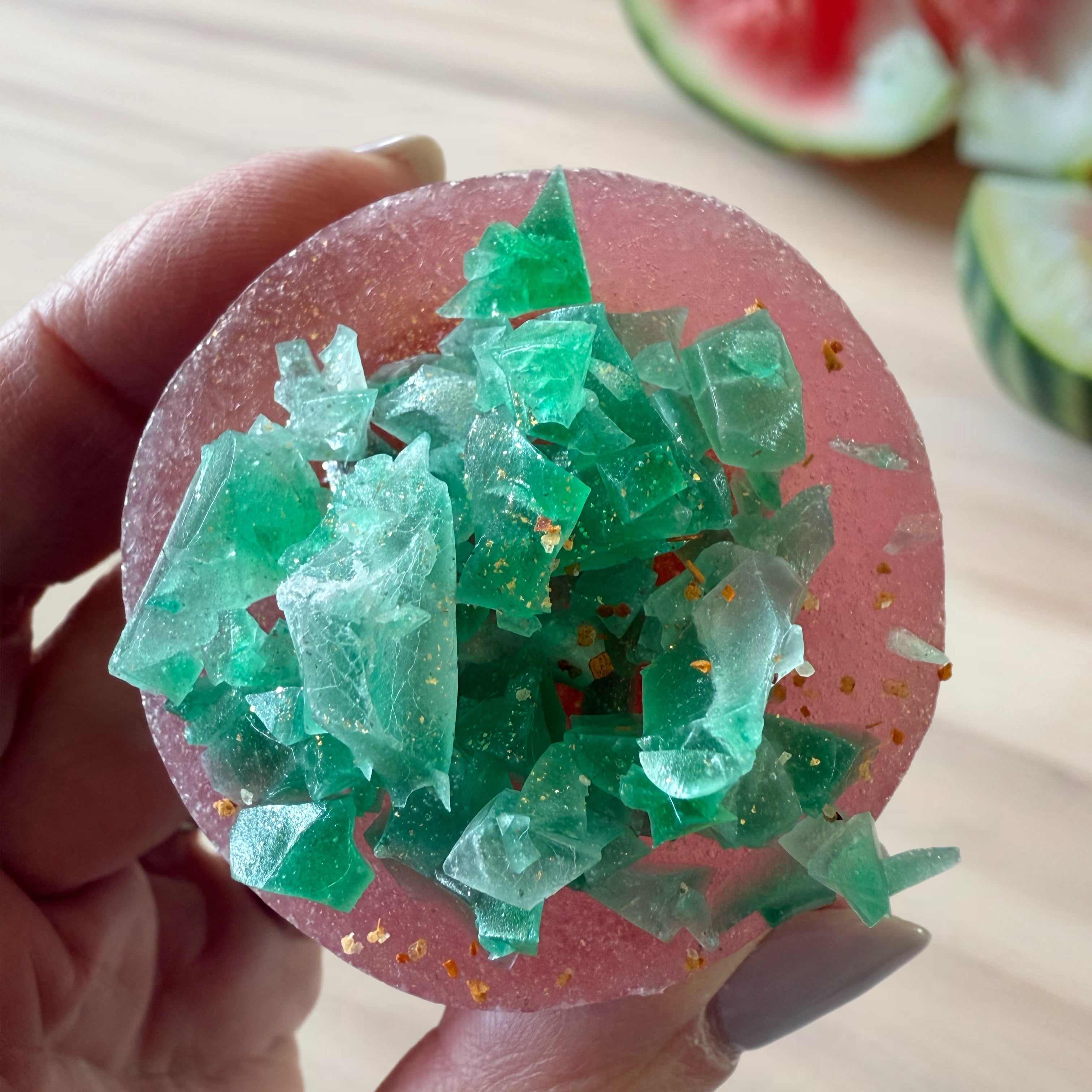 Hand holding a pink and green crystal-like object with watermelon slices in the background