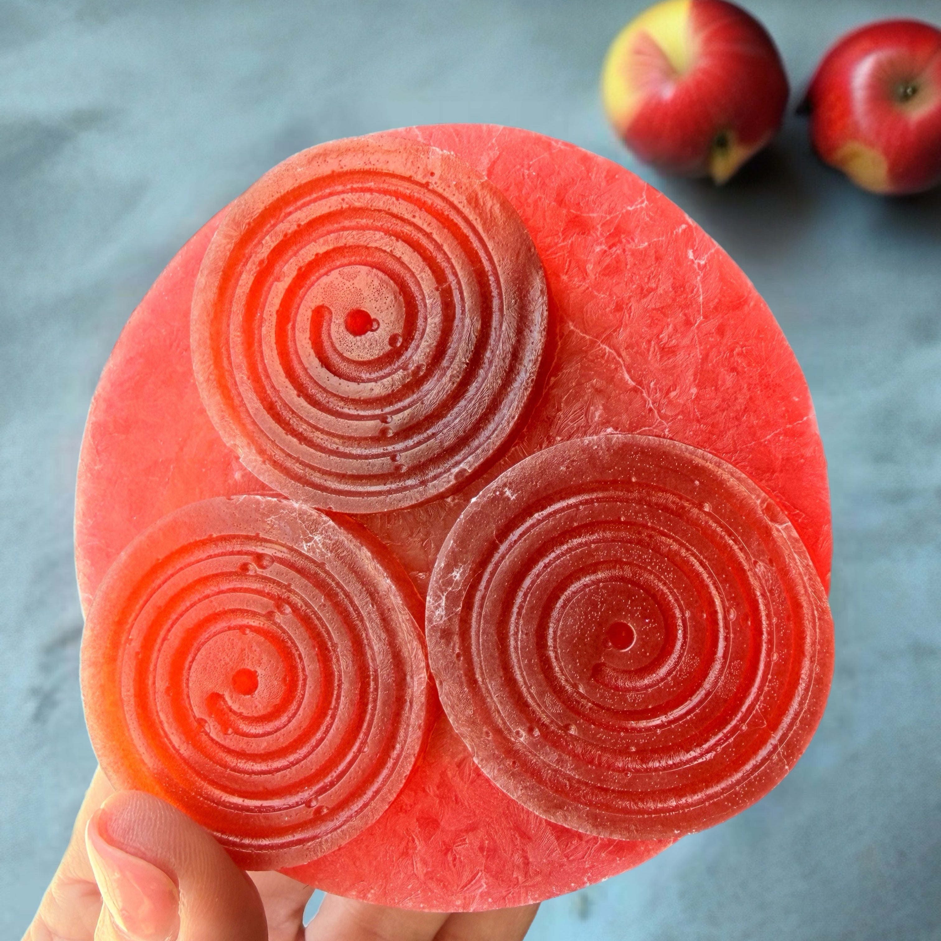 A hand holds a round pink fruit-shaped candy or confection with three spiral swirls on top, with two red apples blurred in the background on a gray surface.